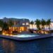 A luxurious floating home is surrounded by calm ocean waters at sunset. The modern structure features large windows, a swimming pool with loungers, and is adorned with vibrant lighting. Tropical palm trees line the house, adding a serene and exotic atmosphere. | MONEY6X