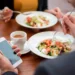 Two individuals are seated at a wooden table, enjoying fresh salads and cups of coffee. One person is holding a fork in one hand and a smartphone in the other, perhaps checking food expenses, while the other is just about to take a bite. The scene suggests a casual, modern dining experience. | MONEY6X