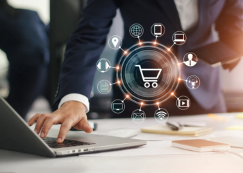 A person in a suit works on a laptop, with a tablet in hand. Various digital icons surround a central shopping cart symbol, illustrating e-commerce concepts such as location, Wi-Fi, email, messaging, and mobile connectivity. | MONEY6X