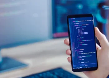 A person holding a smartphone displaying lines of code sits in a workspace emphasizing coding and development activities. The background shows a computer monitor with code, hinting at efforts to build a profitable niche website. Only the person's hand and part of their face are visible. | MONEY6X