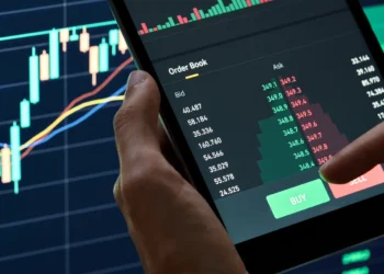 A person uses a tablet displaying live trading data, including a candlestick chart and an order book with bid and ask prices. In the background, a large monitor shows additional financial charts and data, highlighting opportunities for portfolio diversification. The scene depicts an active market trading environment. | MONEY6X