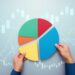 A person's hands arranging a multicolored pie chart against a background featuring financial graphs and stock market candlestick charts, highlighting the importance to diversify your portfolio with segments in green, red, blue, and yellow. | MONEY6X