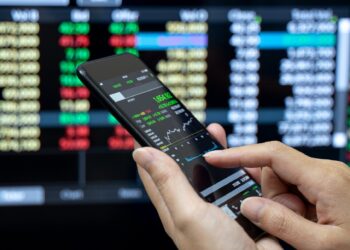 A person uses a smartphone to check financial data. The phone screen shows stock market information, with green and red values, including dividend-paying stocks. In the background, a computer monitor displays various stock charts and figures, indicating active trading or market analysis. | MONEY6X