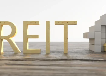 The image displays the word "REITs" formed with golden 3D letters, placed on a wooden surface. To the left of the text is a miniature house, and to the right is a structure made of white cube-shaped blocks with a golden house-shaped block at the top. | MONEY6X