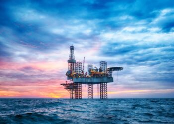 An offshore oil drilling platform stands in the middle of the ocean under a dramatic sunset sky with vibrant pink, purple, and blue hues. The rig's lights are illuminated, and the calm sea reflects the sky's colors, creating a picturesque scene that highlights the allure of investing in commodities. | MONEY6X