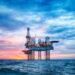 An offshore oil drilling platform stands in the middle of the ocean under a dramatic sunset sky with vibrant pink, purple, and blue hues. The rig's lights are illuminated, and the calm sea reflects the sky's colors, creating a picturesque scene that highlights the allure of investing in commodities. | MONEY6X