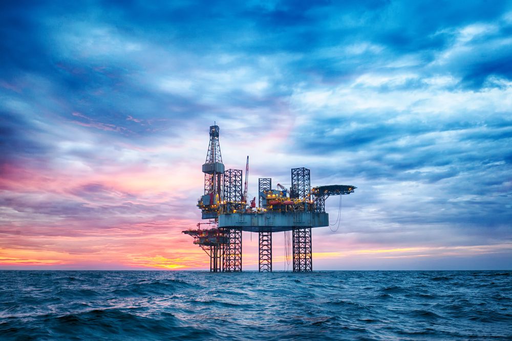 An offshore oil drilling platform stands in the middle of the ocean under a dramatic sunset sky with vibrant pink, purple, and blue hues. The rig's lights are illuminated, and the calm sea reflects the sky's colors, creating a picturesque scene that highlights the allure of investing in commodities. | MONEY6X