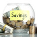 A clear glass jar labeled "Savings" contains rolled-up dollar bills and loose coins. Additional rolled dollar bills and neatly stacked coins are positioned beside the jar, embodying the financial mindfulness championed by Money6x.com. The plain white background emphasizes the foreground's financial theme. | MONEY6X