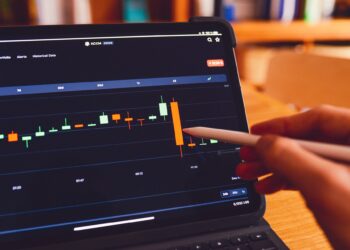 A person using a stylus to interact with a tablet displaying a financial stock chart. The screen shows a candlestick graph with various colors representing market data, alongside information about Certificates of Deposit. The background is blurred, focusing attention on the screen and the stylus. | MONEY6X