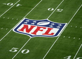 A football field with the NFL logo at the center. The logo features a football and white stars on a blue background at the top, with "NFL" written in white letters on a red background at the bottom. Visible 50-yard lines add to its allure for fans interested in how to make money betting on sports. | MONEY6X