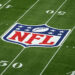 A football field with the NFL logo at the center. The logo features a football and white stars on a blue background at the top, with "NFL" written in white letters on a red background at the bottom. Visible 50-yard lines add to its allure for fans interested in how to make money betting on sports. | MONEY6X