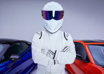 A person wearing a full white racing suit and helmet with a reflective visor stands with arms crossed. Behind them are two sports cars, one blue and one red, in a well-lit studio, demonstrating how you can convey style and charisma without showing your face on platforms like OnlyFans. | MONEY6X