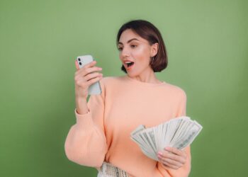 A woman in a peach-colored sweater looks surprised while holding a smartphone in her right hand and a large stack of cash in her left hand, as if she just discovered secret websites to make money. She stands in front of a plain green background. | MONEY6X