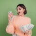 A woman in a peach-colored sweater looks surprised while holding a smartphone in her right hand and a large stack of cash in her left hand, as if she just discovered secret websites to make money. She stands in front of a plain green background. | MONEY6X