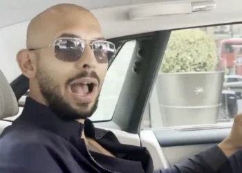 A person with a shaved head and sunglasses is sitting in a car, possibly pondering How Did Andrew Tate Make His Money. They are wearing a dark jacket and have their mouth open as if speaking. The car interior appears luxury with a beige headrest, and there is a cityscape visible through the window. | MONEY6X