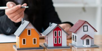 A person holding a pen points toward three miniature house models on a wooden desk. The models, in yellow, red, and white with distinct architectural designs, represent various real estate options. The person is partially visible, focusing intently on the houses. | MONEY6X