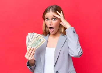 A surprised woman with shoulder-length blonde hair stands against a red background. Holding a fan of U.S. dollar bills in one hand and her other hand on her forehead, she looks amazed, embodying the essence of "How to Make Money Fast as a Woman." She is dressed in a grey blazer over a white shirt. | MONEY6X