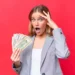 A surprised woman with shoulder-length blonde hair stands against a red background. Holding a fan of U.S. dollar bills in one hand and her other hand on her forehead, she looks amazed, embodying the essence of "How to Make Money Fast as a Woman." She is dressed in a grey blazer over a white shirt. | MONEY6X