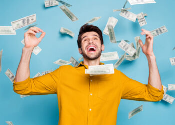 A man in a yellow shirt is standing against a blue background, celebrating joyfully while U.S. dollar bills are falling around him. He has a wide smile and his arms are raised in excitement, reveling in his newfound ability to make money effortlessly. | MONEY6X