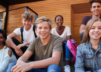 A group of six diverse teenagers, casually dressed, sit on outdoor steps, smiling at the camera. The background features wooden and brick walls. They seem to be discussing how to make money as a teenager while enjoying each other's company. | MONEY6X