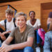 A group of six diverse teenagers, casually dressed, sit on outdoor steps, smiling at the camera. The background features wooden and brick walls. They seem to be discussing how to make money as a teenager while enjoying each other's company. | MONEY6X