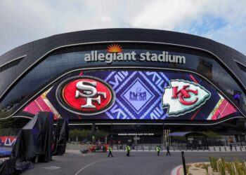 A wide view of Allegiant Stadium with a large electronic display featuring the logos of the San Francisco 49ers and the Kansas City Chiefs, indicating an upcoming game between the two teams. The stadium exterior is sleek and modern with visible walkways and plants—imagine this excitement when you attend Super Bowl 2024. | MONEY6X