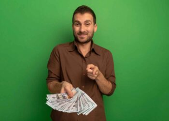 A man with a beard and brown shirt stands in front of a green background, holding a stack of U.S. dollar bills in one hand while pointing towards the camera with the other, suggesting the true meaning of money with a slight smile on his face. | MONEY6X