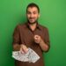 A man with a beard and brown shirt stands in front of a green background, holding a stack of U.S. dollar bills in one hand while pointing towards the camera with the other, suggesting the true meaning of money with a slight smile on his face. | MONEY6X