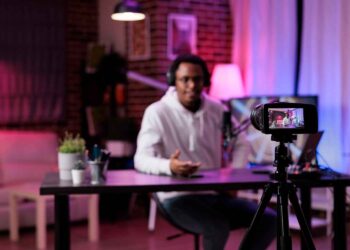 A person is seated at a desk in a warmly lit room, speaking into a microphone about how to make money on YouTube, and wearing headphones. They are being recorded by a camera on a tripod. Various items, including a plant and stationery, are on the desk, with blurred background decor visible. | MONEY6X