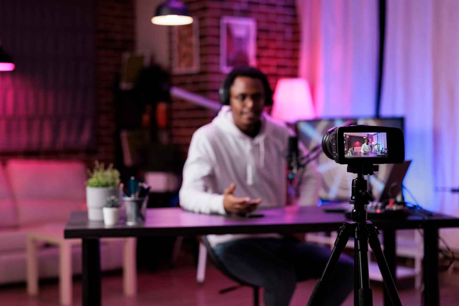 A person is seated at a desk in a warmly lit room, speaking into a microphone about how to make money on YouTube, and wearing headphones. They are being recorded by a camera on a tripod. Various items, including a plant and stationery, are on the desk, with blurred background decor visible. | MONEY6X