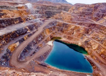 Aerial view of an open-pit mine with terraced, rocky layers. A large, heart-shaped turquoise pool of water sits at the bottom. The winding paths lead down to the pool, and the surrounding terrain is dry and mountainous—an intriguing site for rare earth stocks exploration. | MONEY6X