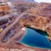 Aerial view of an open-pit mine with terraced, rocky layers. A large, heart-shaped turquoise pool of water sits at the bottom. The winding paths lead down to the pool, and the surrounding terrain is dry and mountainous—an intriguing site for rare earth stocks exploration. | MONEY6X