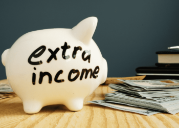 A white piggy bank with "extra income" written on it sits on a wooden table. Nearby, stacks of dollar bills and closed books hint at strategies for saving money and financial growth, subtly suggesting how to make extra money. | MONEY6X