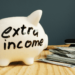A white piggy bank with "extra income" written on it sits on a wooden table. Nearby, stacks of dollar bills and closed books hint at strategies for saving money and financial growth, subtly suggesting how to make extra money. | MONEY6X