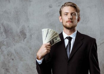 A man wearing a black suit and tie stands against a gray, textured background. He holds several fan-spread U.S. dollar bills in one hand near his face, looking directly at the camera with a neutral expression, as if contemplating ways to invest money. | MONEY6X