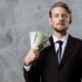A man wearing a black suit and tie stands against a gray, textured background. He holds several fan-spread U.S. dollar bills in one hand near his face, looking directly at the camera with a neutral expression, as if contemplating ways to invest money. | MONEY6X