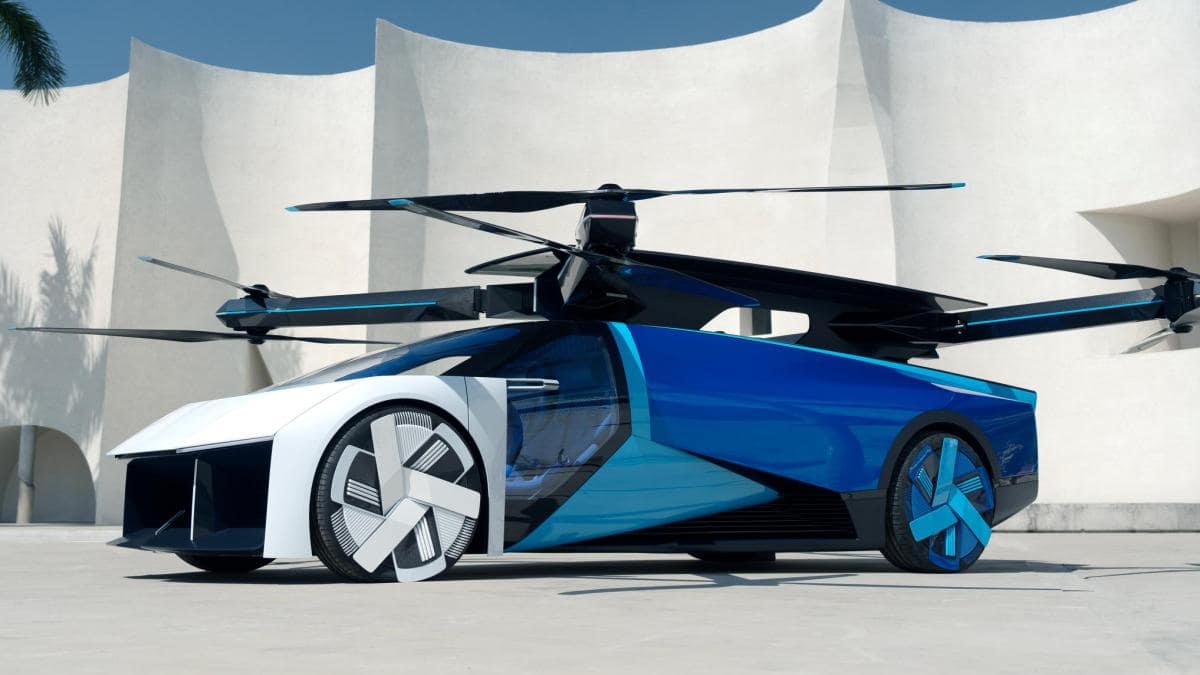 A sleek futuristic blue and white flying car with four large propellers on top, parked in front of a modern white building with curved architectural elements. Investors eyeing flying car stocks are excited by its streamlined design and large wheels, combining features of both a car and a drone. | MONEY6X