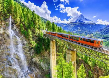 A bright orange train travels across a tall, narrow bridge surrounded by lush greenery and pine trees. A cascading waterfall flows down the cliffside on the left. Snow-capped mountains rise in the background under a bright, cloud-filled blue sky, capturing the essence of Luxury Swiss Vacations. | MONEY6X