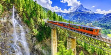 A bright orange train travels across a tall, narrow bridge surrounded by lush greenery and pine trees. A cascading waterfall flows down the cliffside on the left. Snow-capped mountains rise in the background under a bright, cloud-filled blue sky, capturing the essence of Luxury Swiss Vacations. | MONEY6X