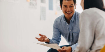 A man in a blue shirt is sitting and smiling while holding a paper, engaging in a conversation with a woman with long dark hair who is facing away from the camera. They are in a well-lit room with blurred documents and notes on the wall, possibly discussing hiring details. | MONEY6X