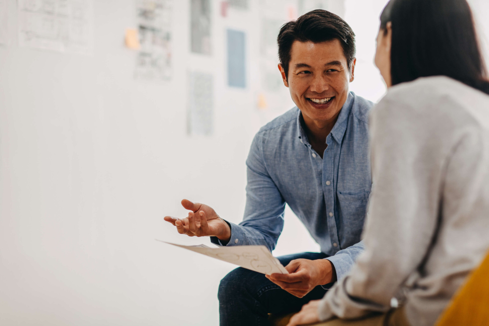 A man in a blue shirt is sitting and smiling while holding a paper, engaging in a conversation with a woman with long dark hair who is facing away from the camera. They are in a well-lit room with blurred documents and notes on the wall, possibly discussing hiring details. | MONEY6X