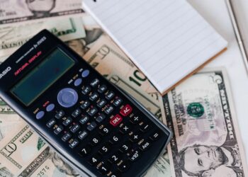 A scientific calculator rests on spread-out US dollar bills, including ten, twenty, and five-dollar notes. To the right of the calculator is a small notepad with a pen placed on top of it, possibly tallying up expenses or calculating a strategy for improving that crucial credit score. | MONEY6X