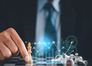 A person in a suit moves a golden chess piece on a chessboard. Silver pieces are toppled over. A transparent graph with rising arrows and bar charts overlays the image, suggesting strategic planning or financial growth, much like the careful strategy required in investing. | MONEY6X