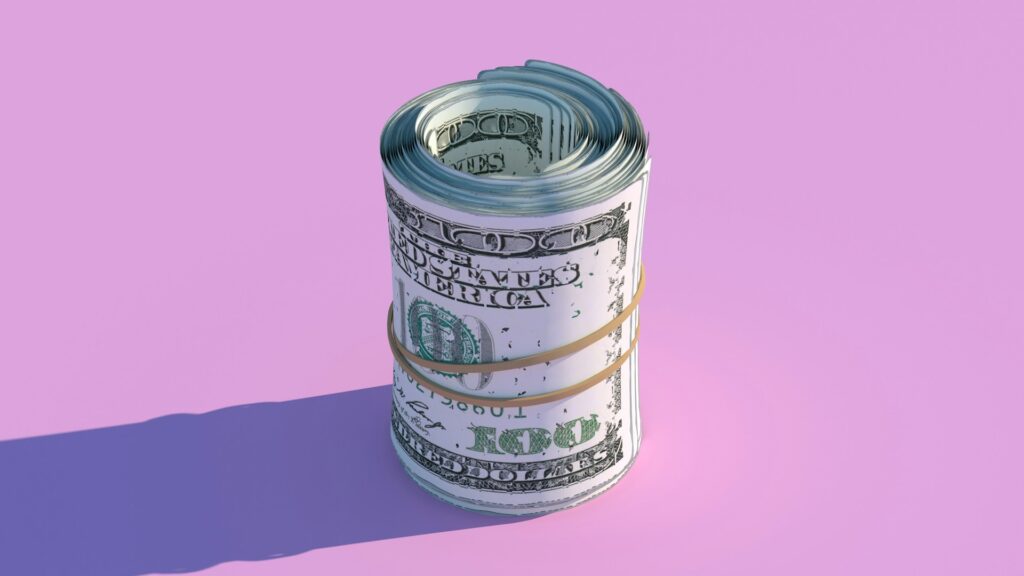 A neatly rolled bundle of U.S. hundred-dollar bills, a symbol of debt management, is secured with a rubber band and positioned upright against a pastel pink background. The roll casts a shadow to the left, adding depth to the image. | MONEY6X