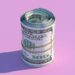 A neatly rolled bundle of U.S. hundred-dollar bills, a symbol of debt management, is secured with a rubber band and positioned upright against a pastel pink background. The roll casts a shadow to the left, adding depth to the image. | MONEY6X