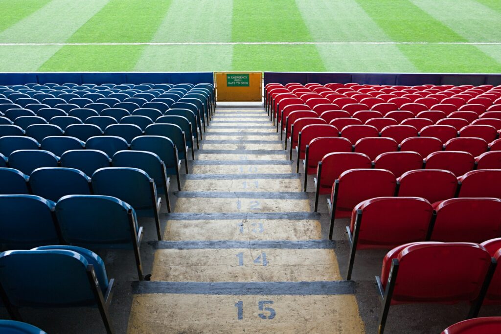 A stadium seating area with blue seats on the left and red seats on the right. The steps between the seats are numbered, leading down to a green field. The neatly mowed grass invites investment in breathtaking events. A yellow exit sign is visible at the bottom. | MONEY6X