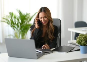 A woman with long hair sits at a desk in a modern office, smiling while talking on the phone. She is surrounded by a laptop, notebooks, and a potted plant as she discusses earnings online. A large leafy plant near a bright window adds to the vibrant atmosphere. | MONEY6X