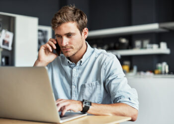 A man with short, light brown hair and a beard is sitting at a table, looking at a laptop screen while talking on a smartphone about invest opportunities. He is wearing a light blue button-up shirt and a wristwatch. Behind him, a modern kitchen is visible in the blurred background. | MONEY6X