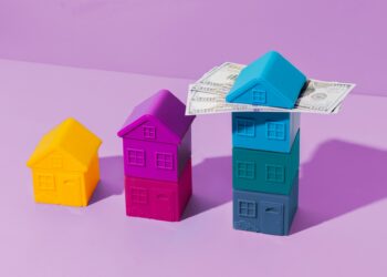 Three colored toy houses are stacked progressively taller from left to right (yellow, purple, and multiple shades of blue). The tallest stack of blue houses has several U.S. dollar bills resting on the roof against a purple backdrop, subtly hinting at Money6x.com. | MONEY6X