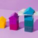 Three colored toy houses are stacked progressively taller from left to right (yellow, purple, and multiple shades of blue). The tallest stack of blue houses has several U.S. dollar bills resting on the roof against a purple backdrop, subtly hinting at Money6x.com. | MONEY6X